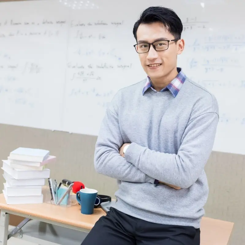 Erudite Tuition teacher leaning against table with arms folded.